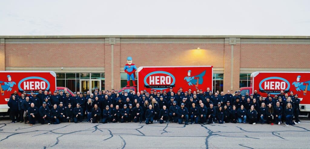 A large group of people in dark clothing poses in front of two red Hero-branded trucks outside a brick building in Bloomington.