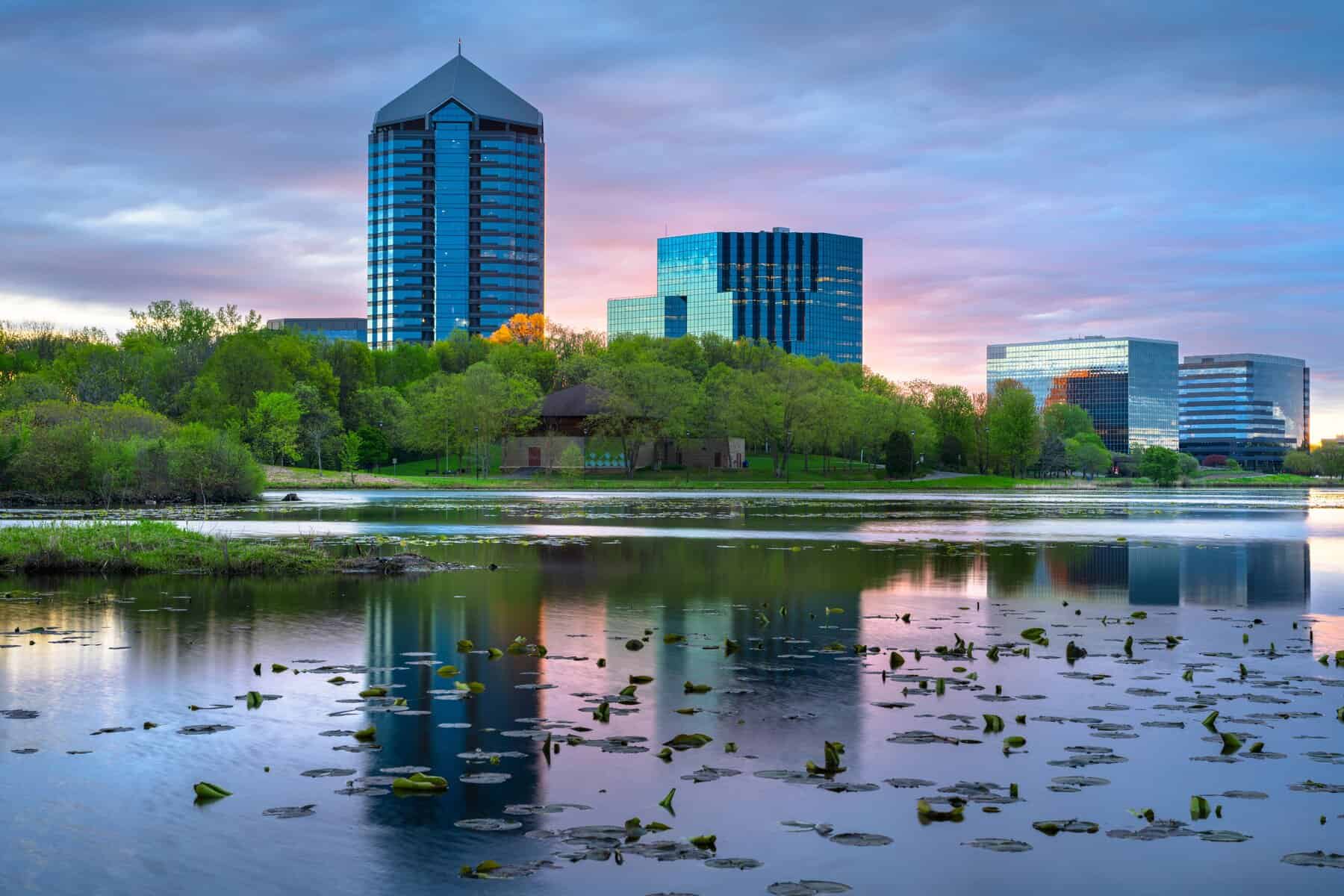 Modern office buildings rise above a tree-lined lakeshore in Bloomington at sunset, with lily pads floating on the calm water in the foreground.