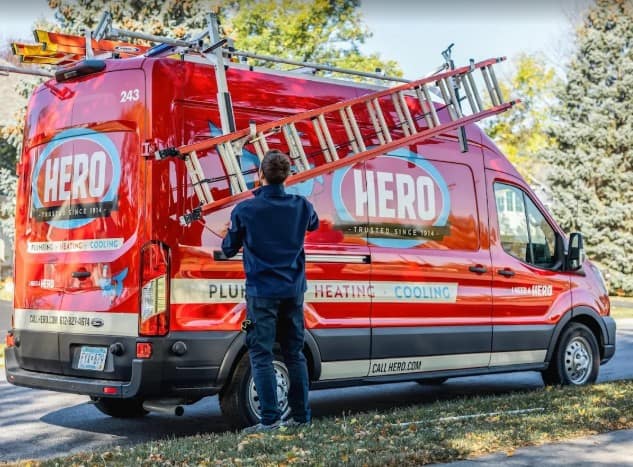 A person in uniform lifts a ladder onto a red HERO plumbing, heating, and cooling service van parked on a Bloomington residential street.