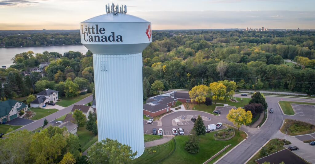 A tall water tower labeled "Little Canada" stands among trees and buildings in a suburban area near a lake, not far from Bloomington.