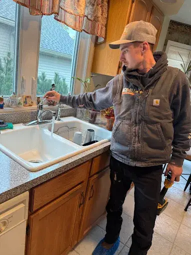 A man in work clothes stands by a kitchen sink, turning on the faucet while holding a tool in his other hand.