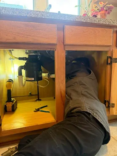 A person is lying on the floor and working under a kitchen sink cabinet with tools and a drill nearby.