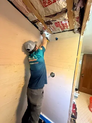 A person in a cap installs insulation and wiring in the ceiling of a partially finished room with bare walls.