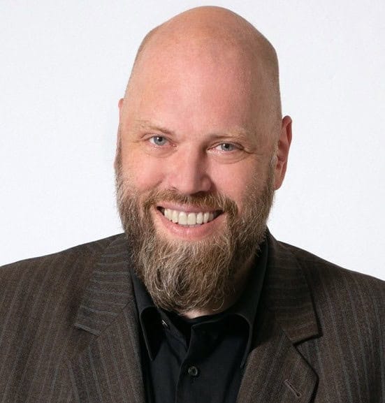 Mann mit Bart und Anzug, lächelnd, vor weißem Hintergrund.