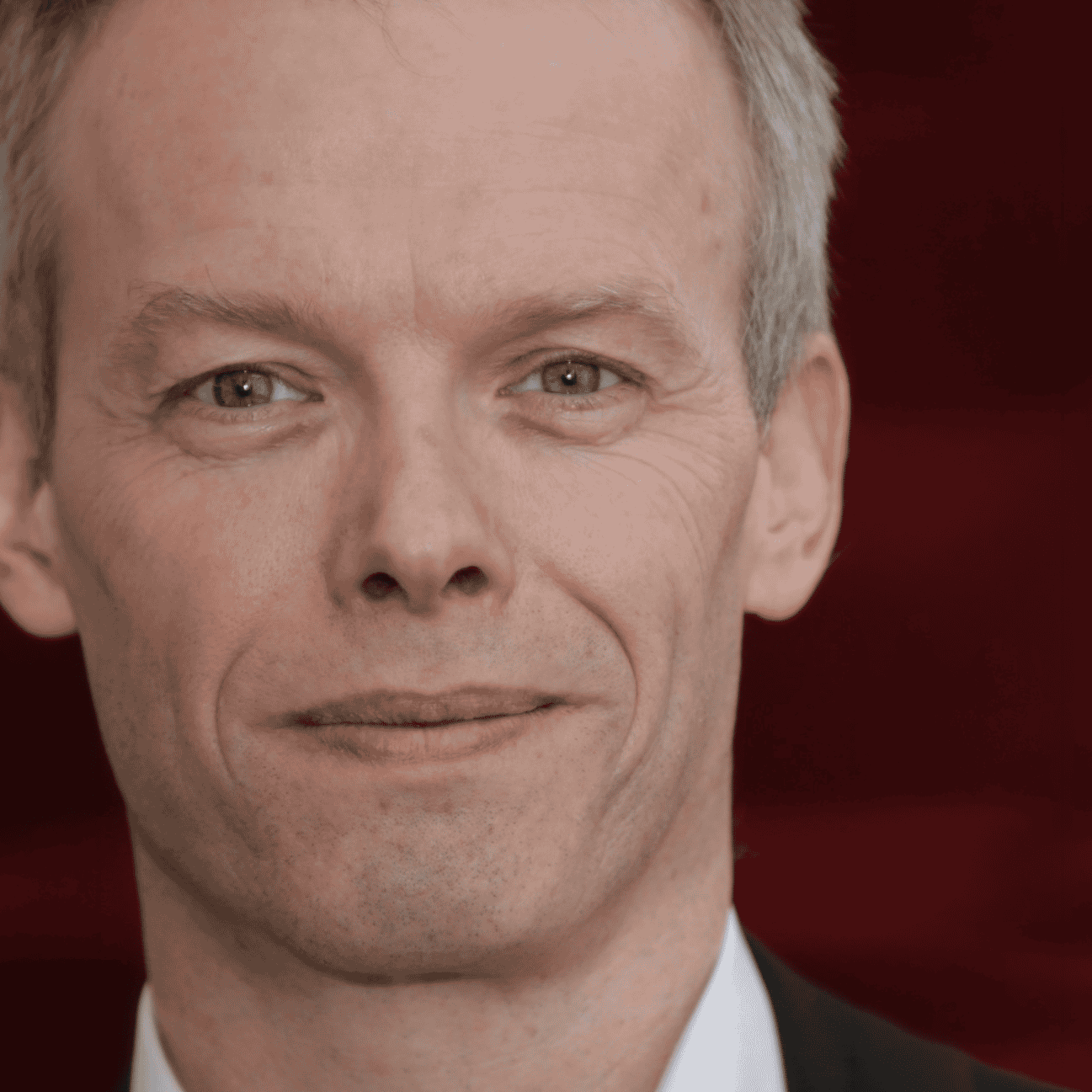 Mann in formeller Kleidung vor rotem Hintergrund, lächelnd.