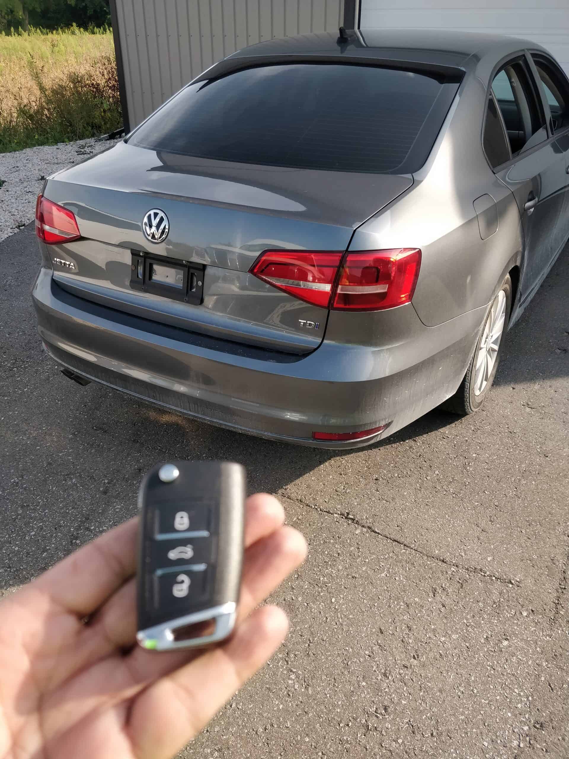 Remote car key fob with lock, unlock, and trunk buttons in front of a parked gray Volkswagen Jetta.