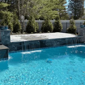 Backyard swimming pool with waterfall and stone deck, surrounded by lush trees and privacy fence.