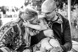 Twee ouders knuffelen hun peuter. De peuter houdt haar moeder vast terwijl moeder en kind elkaar kussen. Een intiem moment uit een Day in the Life vastgelegd door documentair fotograaf Michella Kuijkhoven