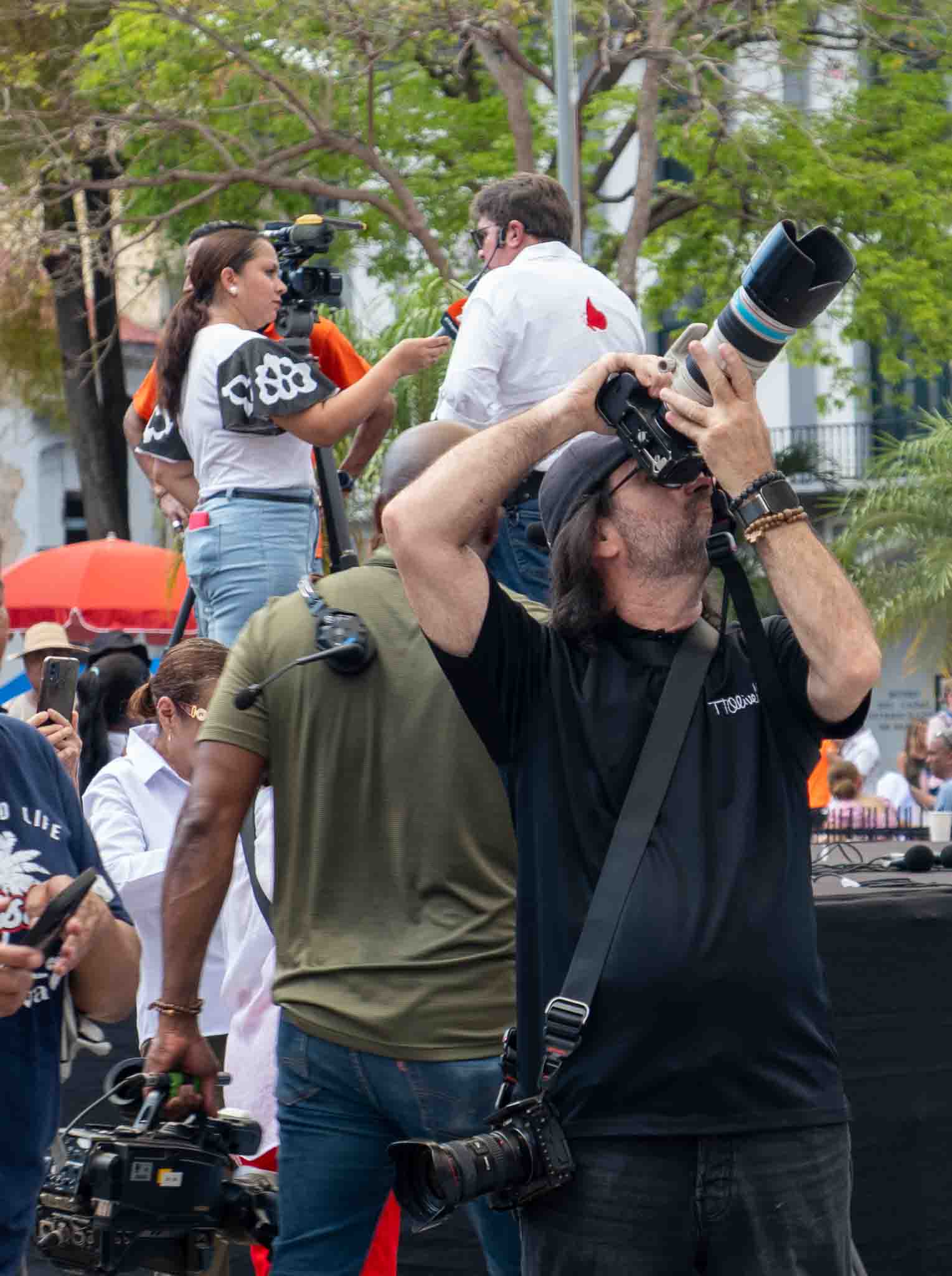 Photographer in black shirt capturing overhead shots with a telephoto lens during a live event in Casco Viejo, surrounded by a media crew