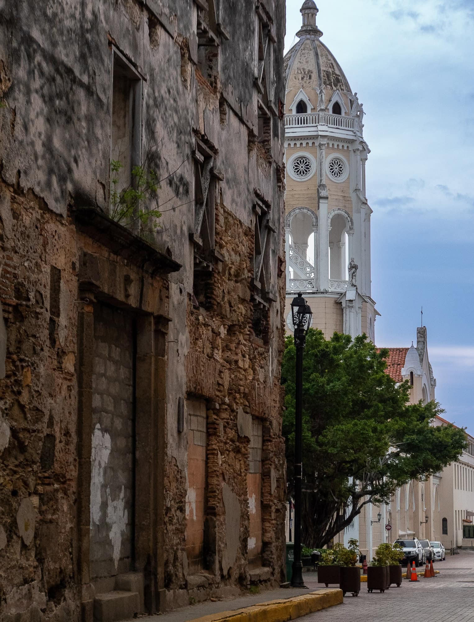Contrast between a decaying colonial facade and the white bell tower of the Metropolitan Cathedral in Casco Viejo, Panama, with cobblestone street and trees below.