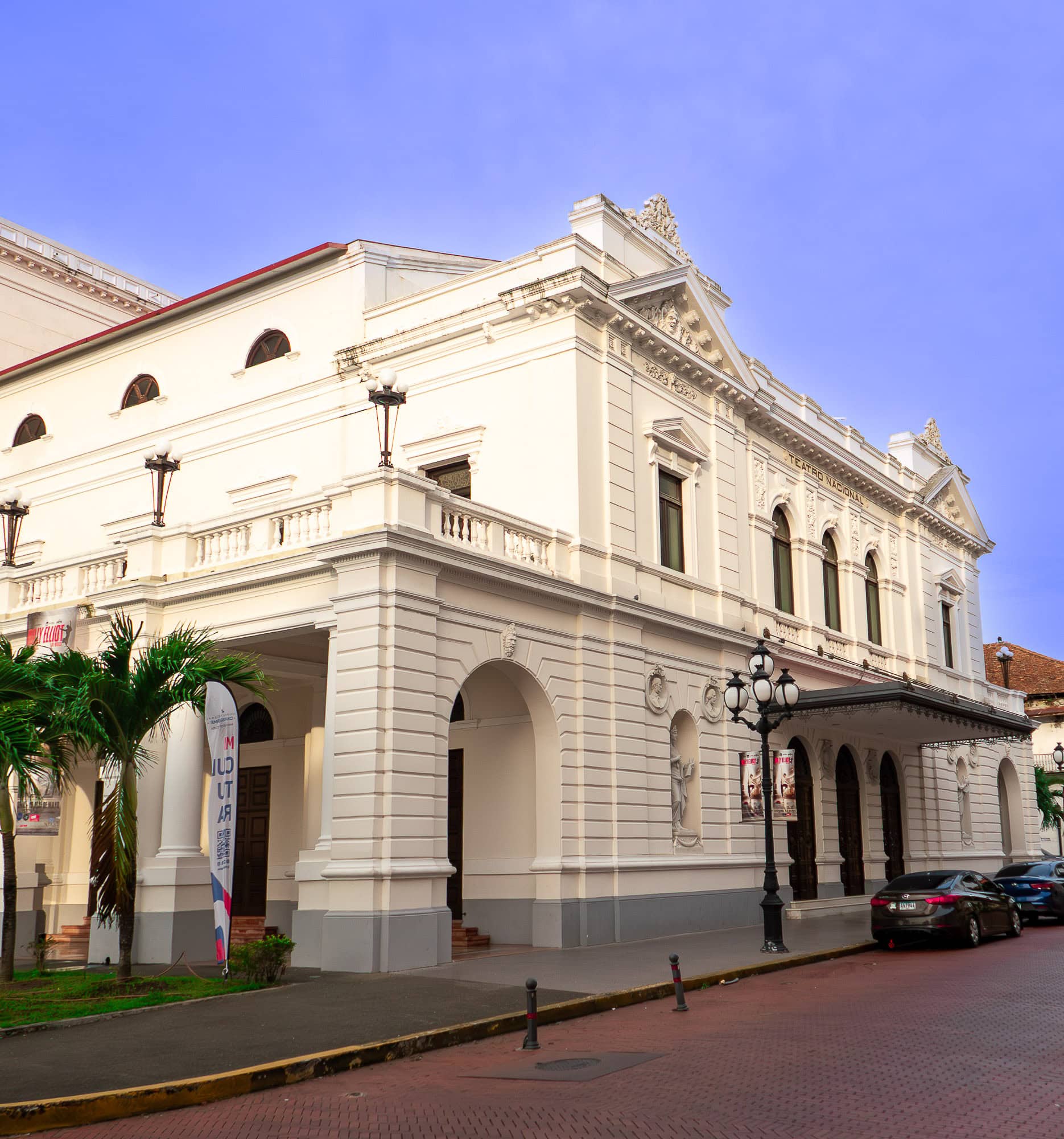Front facade of Teatro Nacional in Casco Viejo, Panama, showing neoclassical columns, arched doorways, decorative statues, and ornate wrought-iron street lamps.