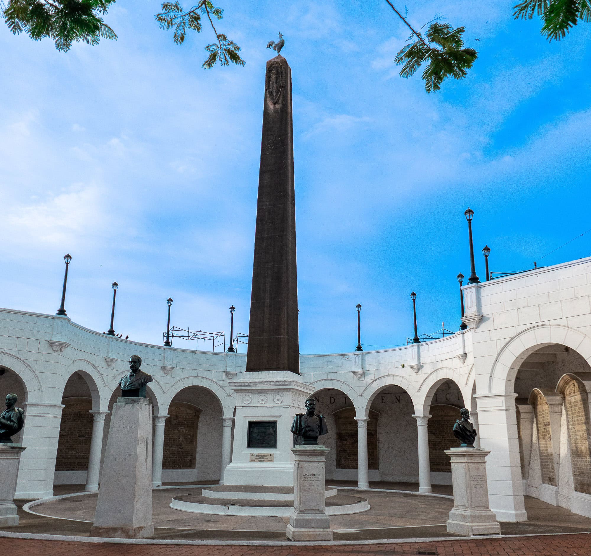 Panoramic shot of the Independence Monument in Plaza de la Independencia, Casco Viejo, featuring statues of national heroes surrounding the central obelisk under a blue sky.