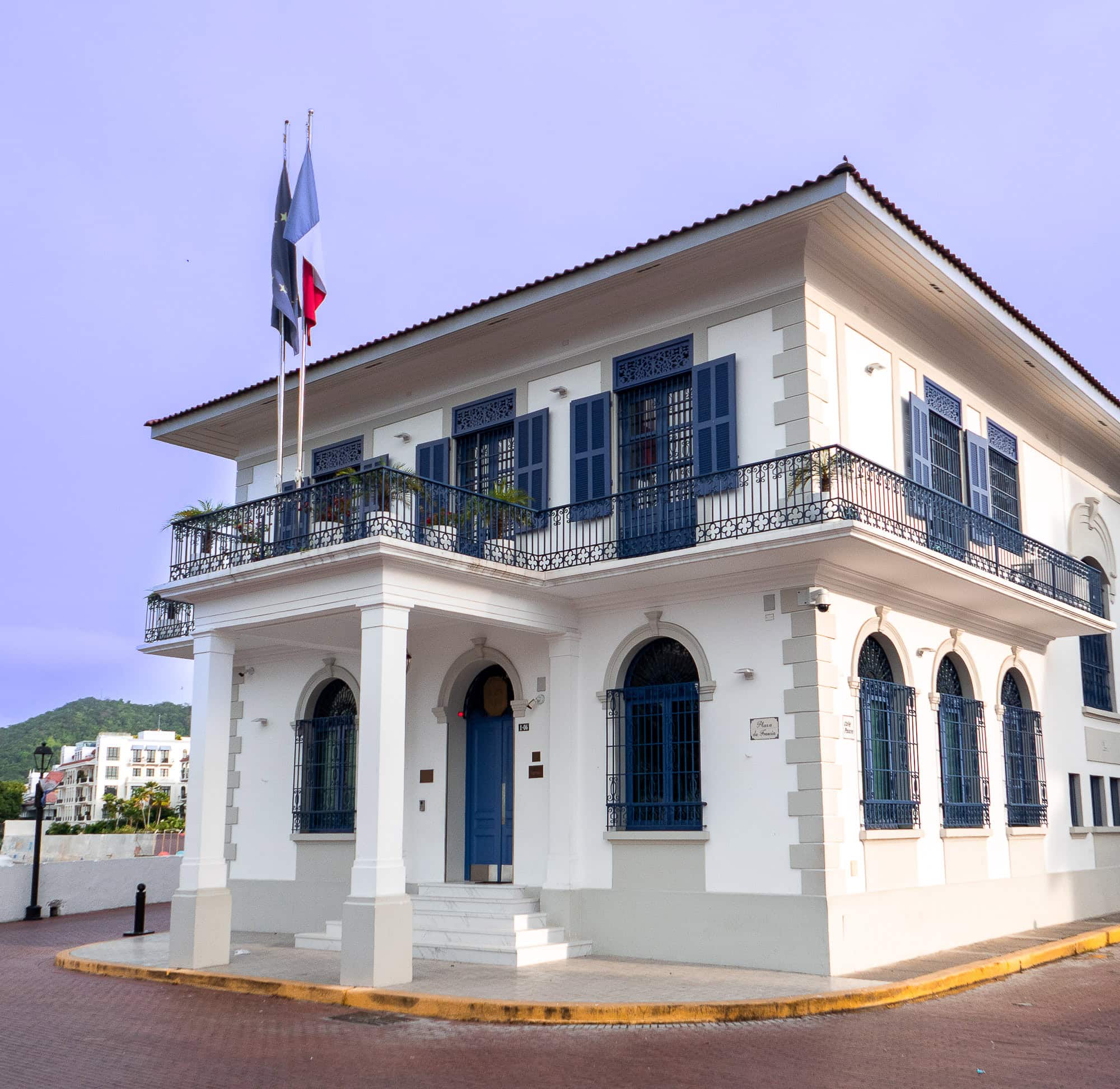 Elegant white building of the French Embassy in Casco Viejo, Panama, with blue shutters and French and Panamanian flags flying above the main entrance.