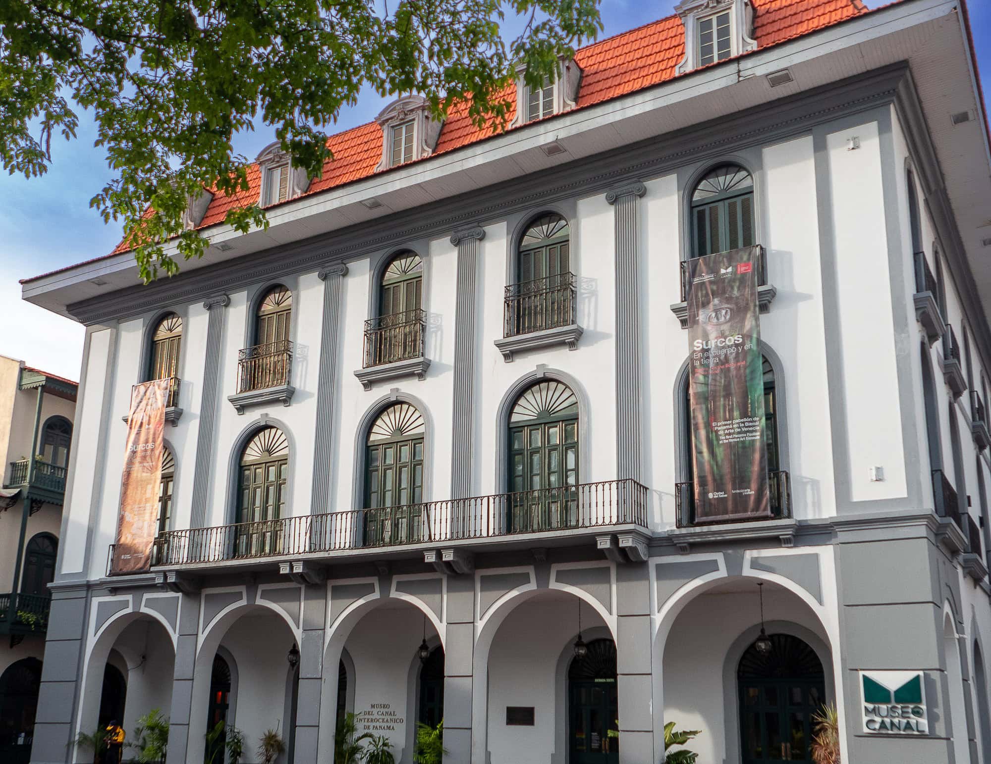 Front view of Museo del Canal Interoceánico in Casco Viejo, Panama, with colonial arches, tall green windows, and cultural banners on display.