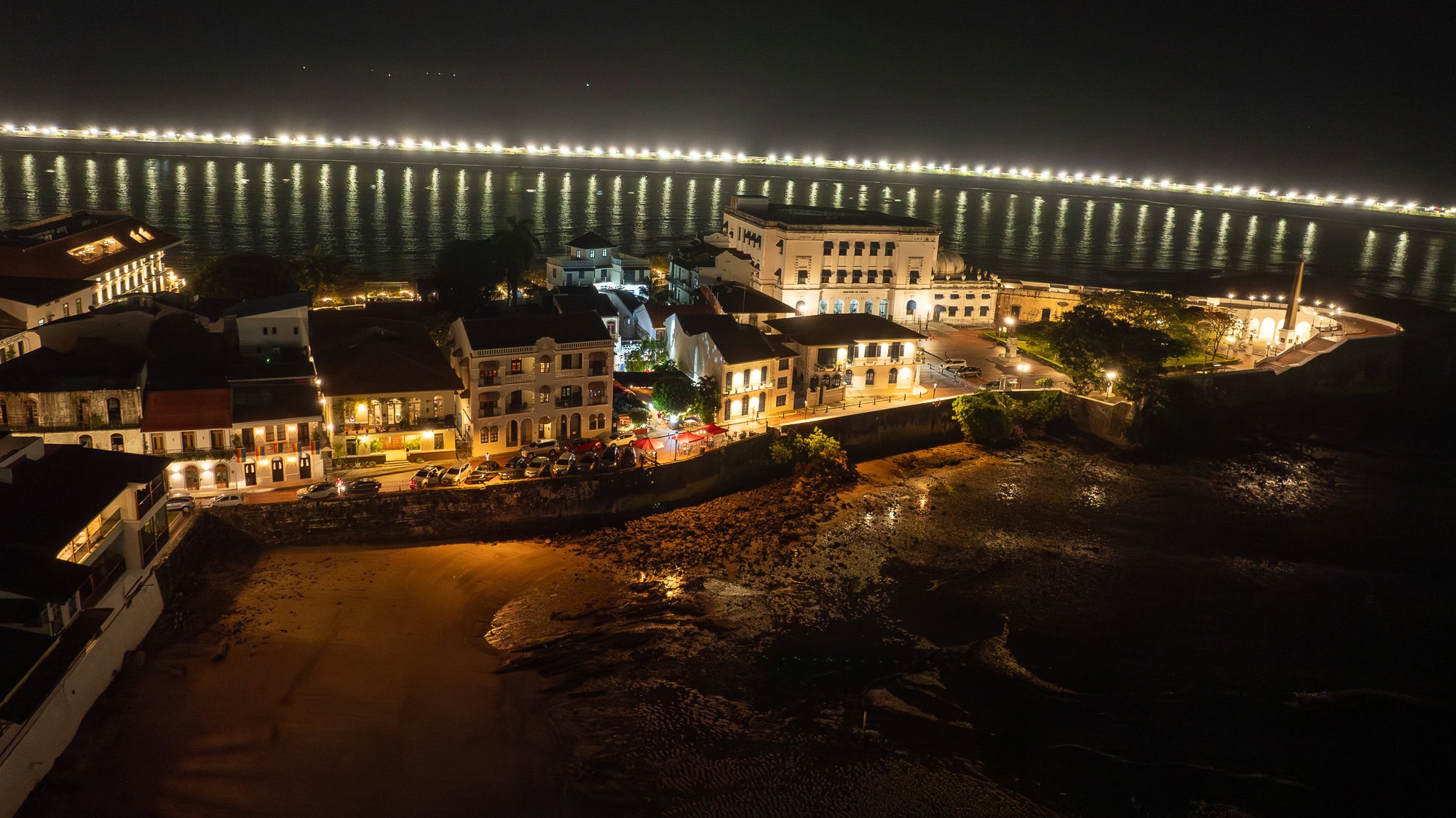 Nighttime aerial view of colonial rooftops and streetlights along the waterfront, with the Pacific coast lit in the distance