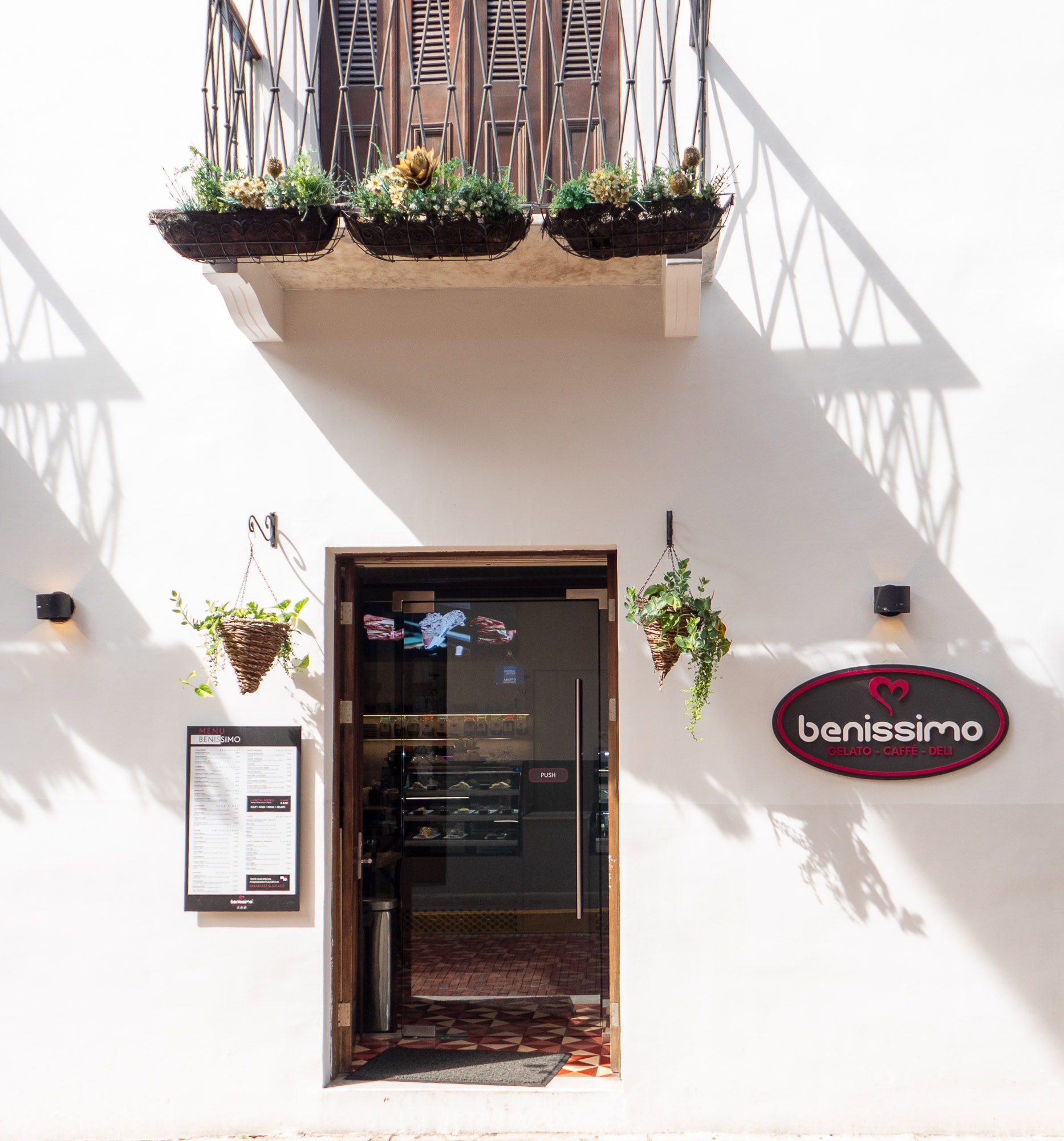 Front entrance of Benissimo Gelato Café + Deli in Casco Viejo, Panama, featuring a menu board, potted plants, and a decorative balcony above the door.