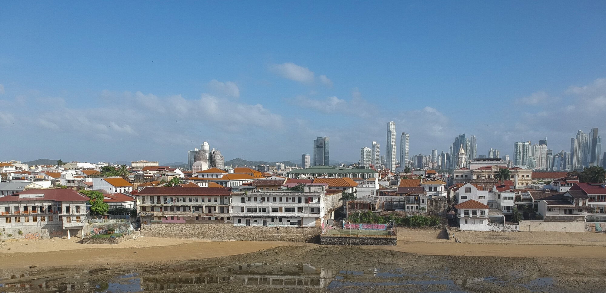 A panoramic view of Casco Viejo’s coastline with historic buildings in the foreground and Panama City’s skyline in the background under a clear blue sky.