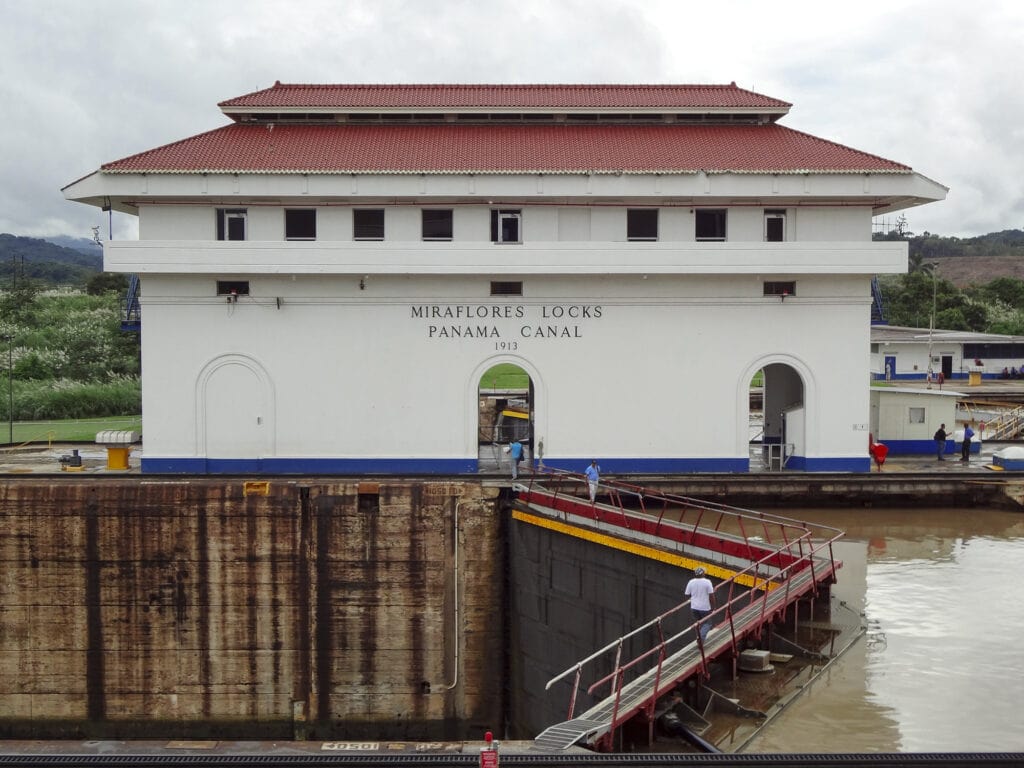 The historic Mira Flores Locks building at the Panama Canal, with canal workers visible near the water’s edge and lock gates.