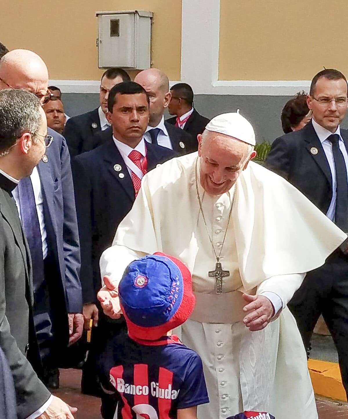 Pope Francis warmly greets a child wearing a red and blue cap during a visit to Casco Viejo, Panama, surrounded by security and local officials.