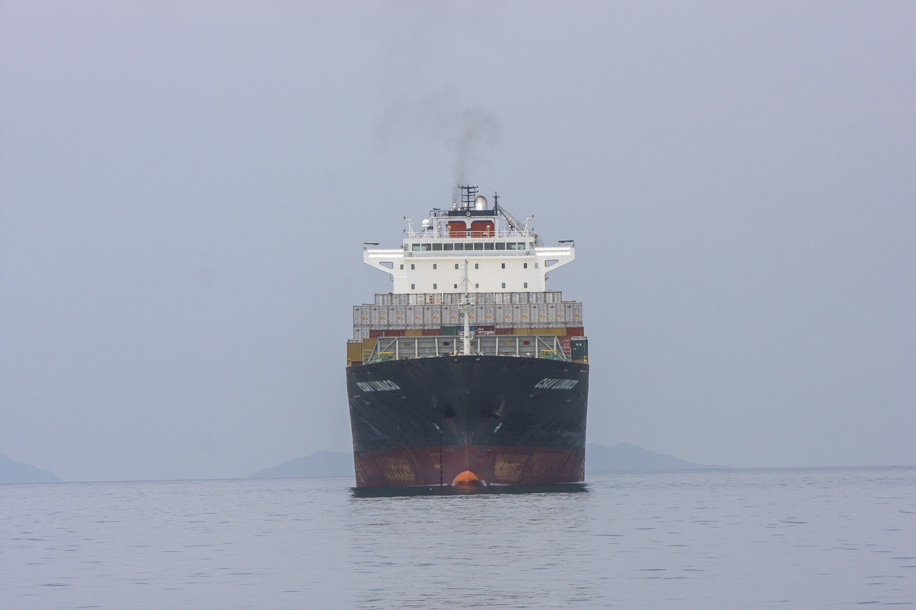 A large cargo ship heads directly toward the camera, preparing to enter the Panama Canal under overcast skies.