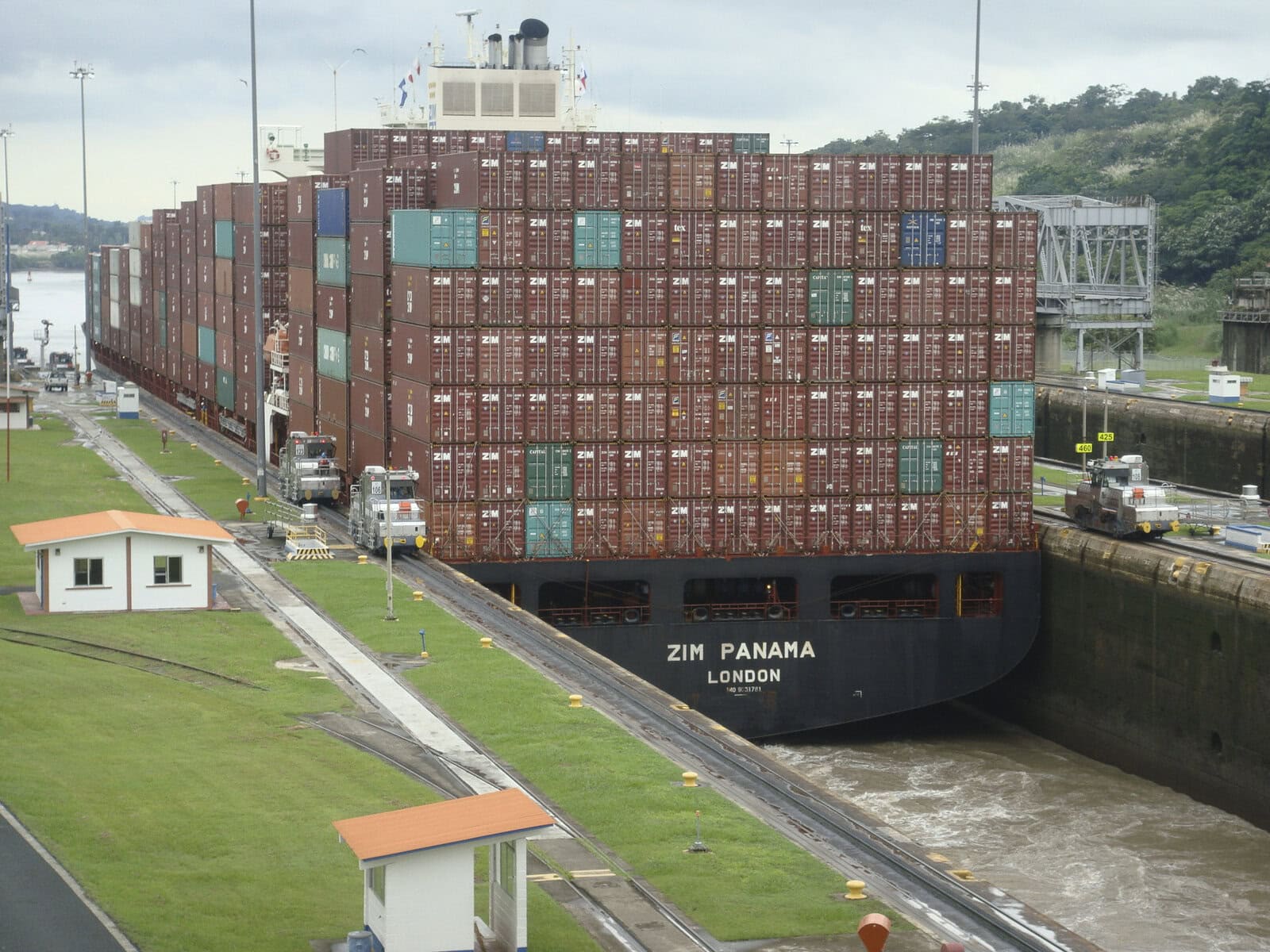 A container ship named ZIM Panama navigates the narrow Mira Flores Locks of the Panama Canal, guided by locomotives on either side.