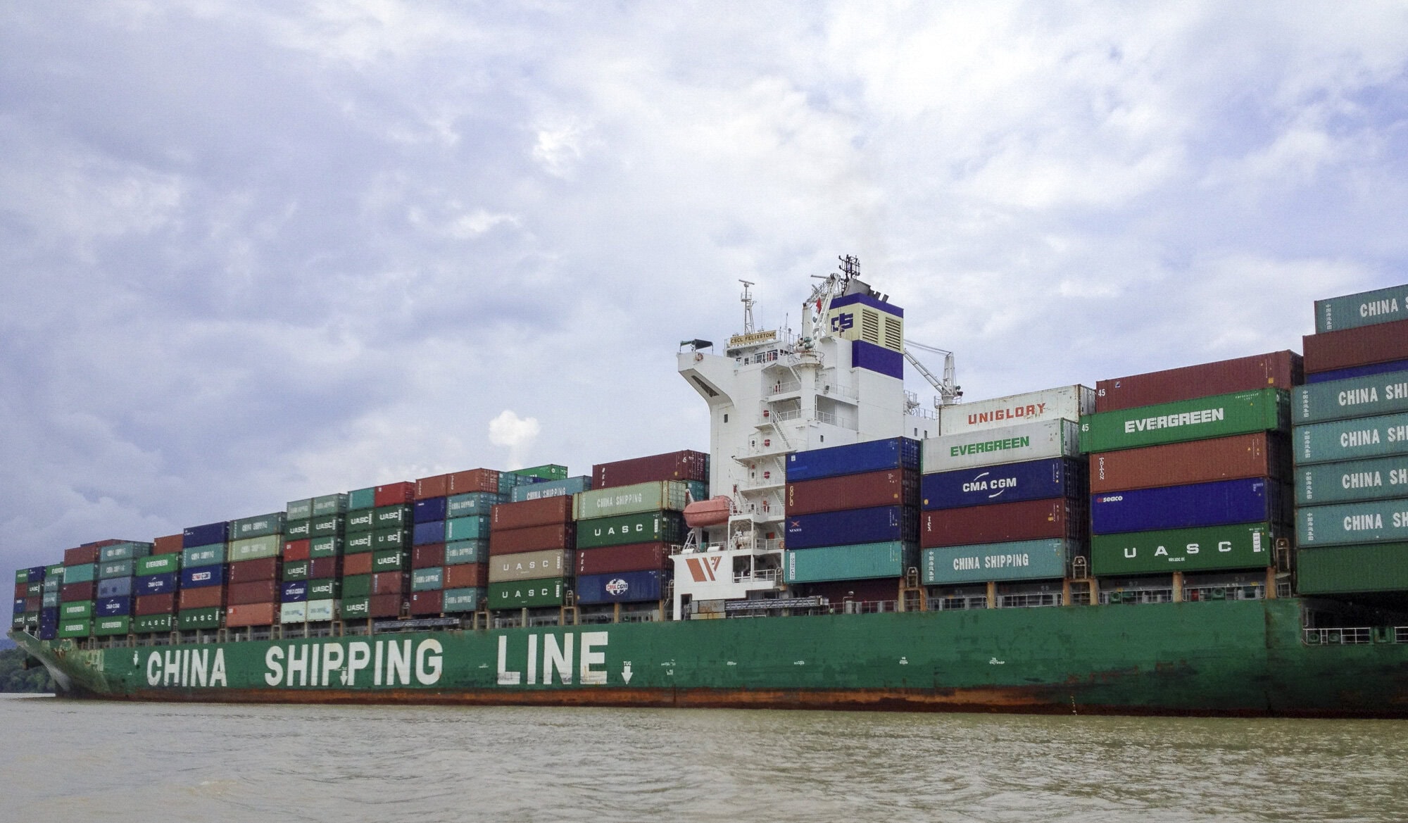 A massive container ship from China Shipping Line transits the Panama Canal, stacked high with colorful cargo containers under a cloudy sky.