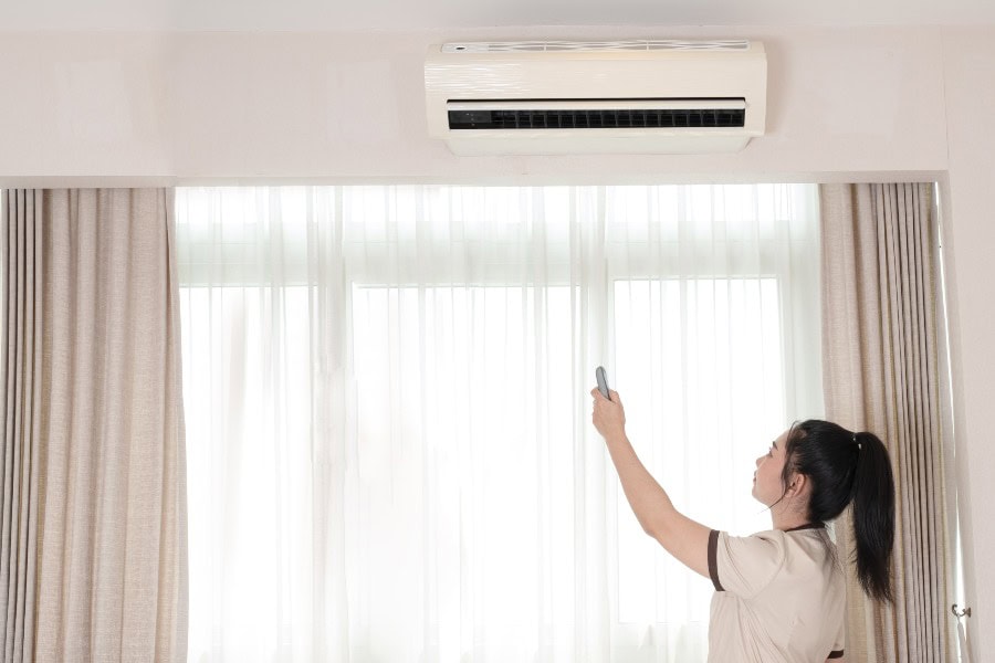 Woman using remote to operate a wall-mounted air conditioner in a bright room.