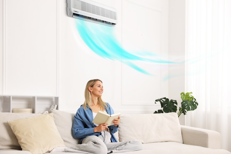 Woman reads book on sofa enjoying cool air flow from active wall AC unit.