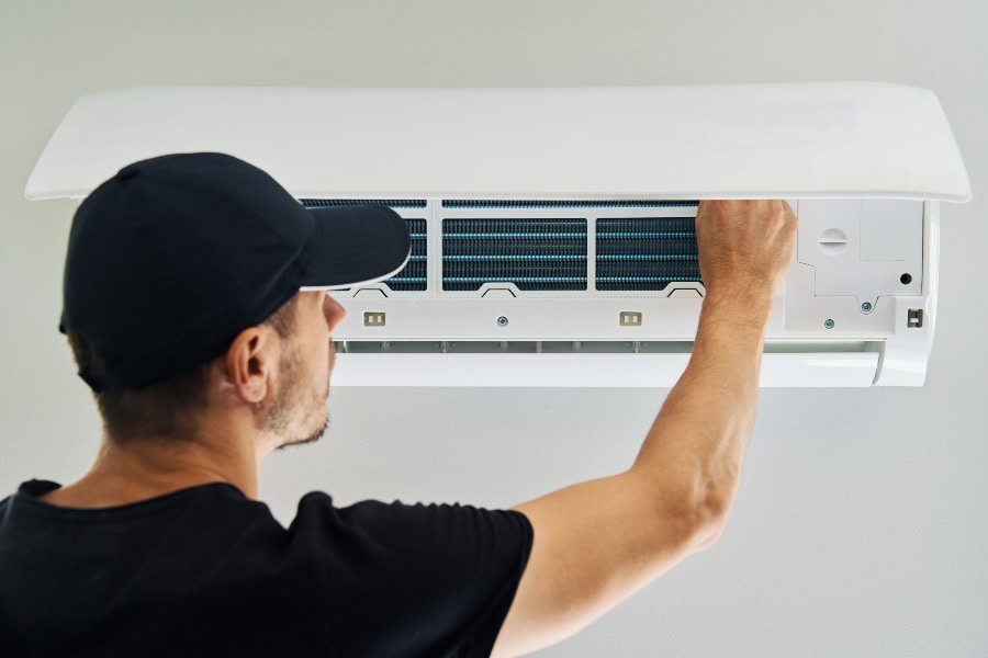 Technician in black cap and shirt opens panel on white wall-mounted air conditioner for maintenance or reset.