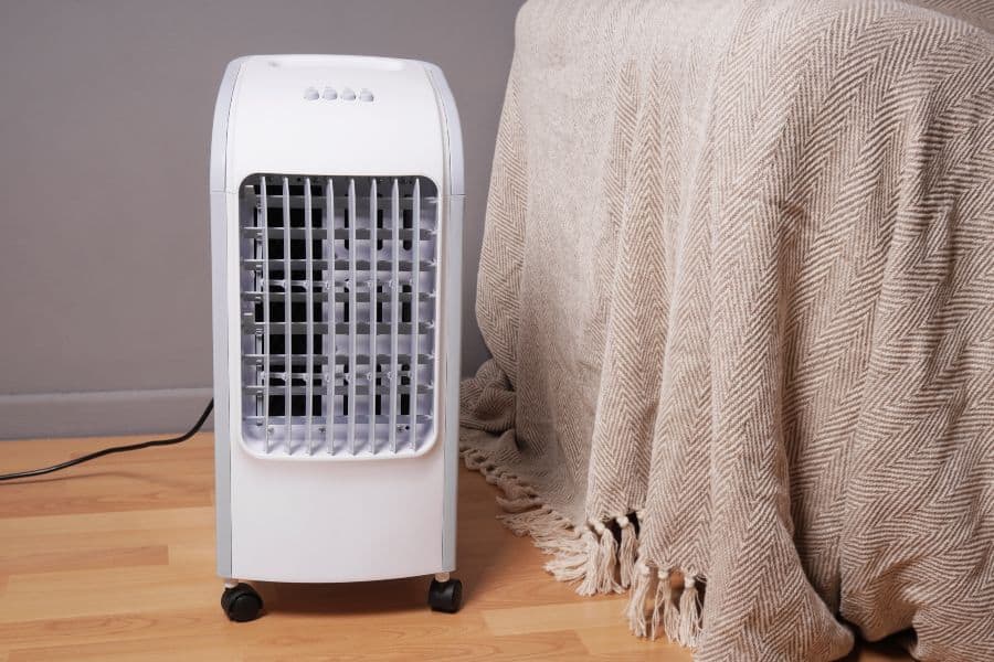 White portable evaporative cooler on wheels near draped bed for AC running costs.