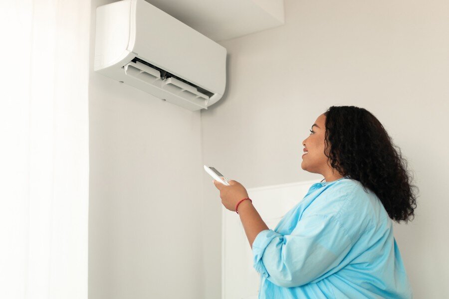 Woman in blue blouse aims remote at white wall AC unit, promoting proactive use to extend lifespan.