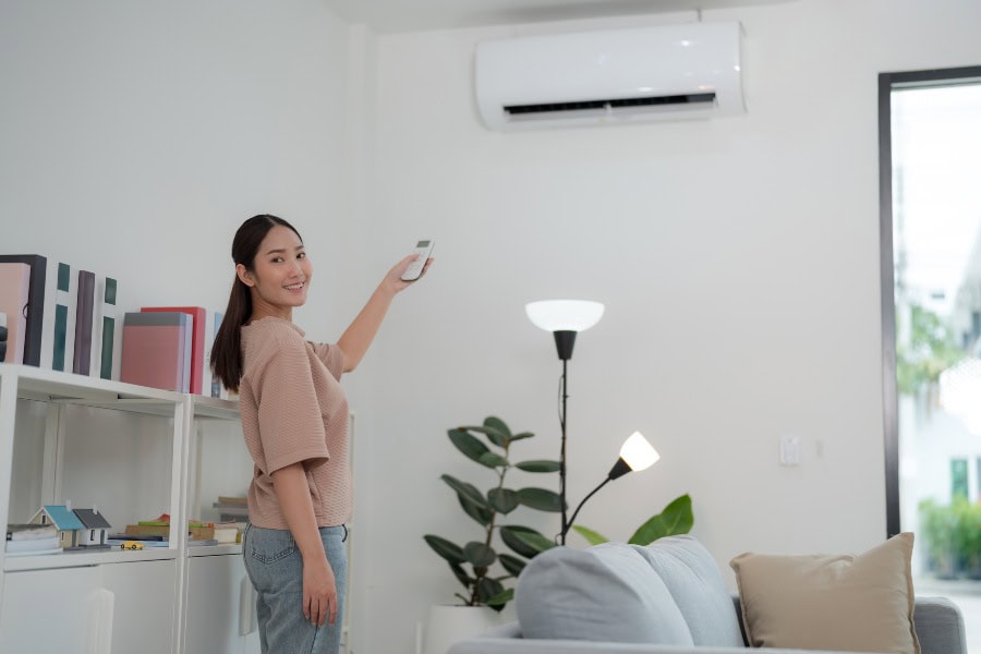 Young woman in beige top points remote at white wall AC unit in modern living room, resetting for peak performance.