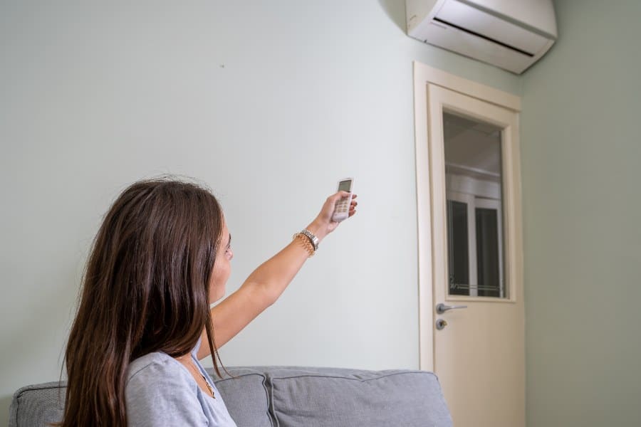 Person activating air conditioner with remote from couch, illustrating heatwave expenses.