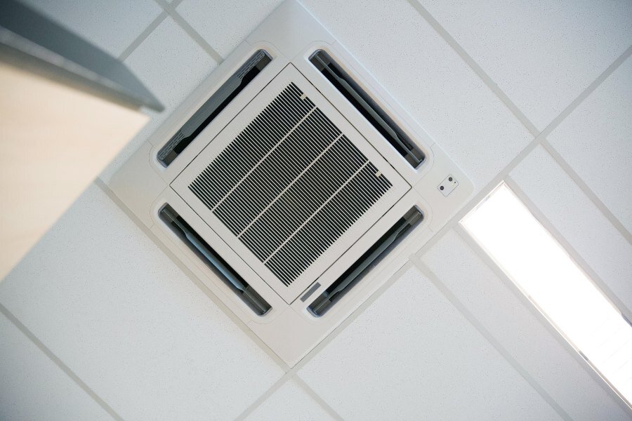 Ceiling-mounted air conditioner in a tiled office ceiling beside a bright fluorescent light.