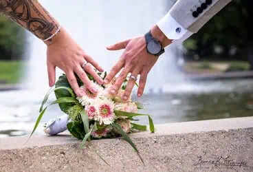 Blumenstrauß und Hände eines Paares, Hochzeitsfotografie in Aachen, verliebtes Paar mit Ringen und eleganten Outfits, romantische Kulisse.