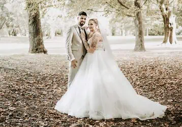 Hochzeitsfoto eines Paares im Park mit Baum Hintergrund, Braut im weißen Kleid, Bräutigam in hellgrauem Anzug, romantische Stimmung.