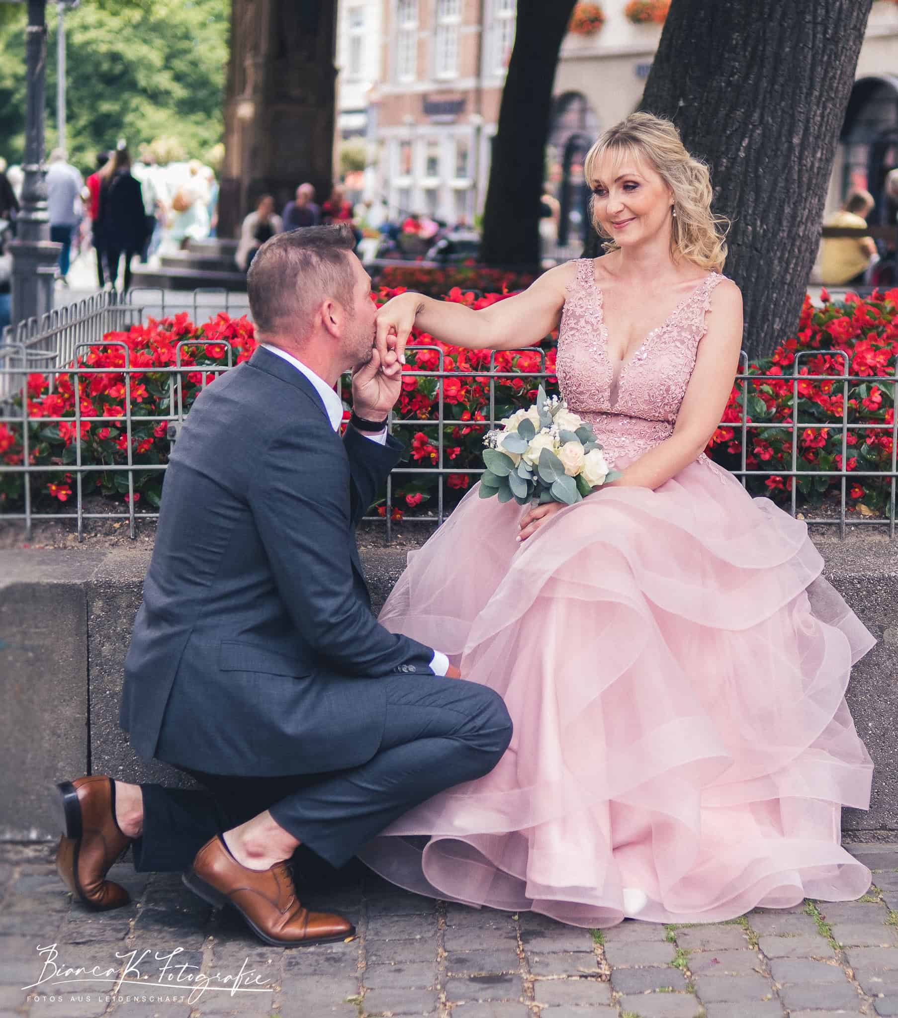 Blickfang: Romantisches Hochzeitsfoto von einem Paar in Aachen, Frau in eleganter rosa Brautkleidung, Mann im Anzug, liebevolle Geste.