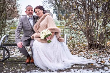 Hochzeitsfoto im Winter, Braut und Bräutigam sitzen auf einer Parkbank, unter Bäumen mit frostigem Hintergrund, bei einer Hochzeit in Aachen.