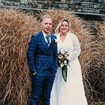 Hochzeitspaar in eleganter Kleidung vor natürlichen Pflanzen und Steinmauer.