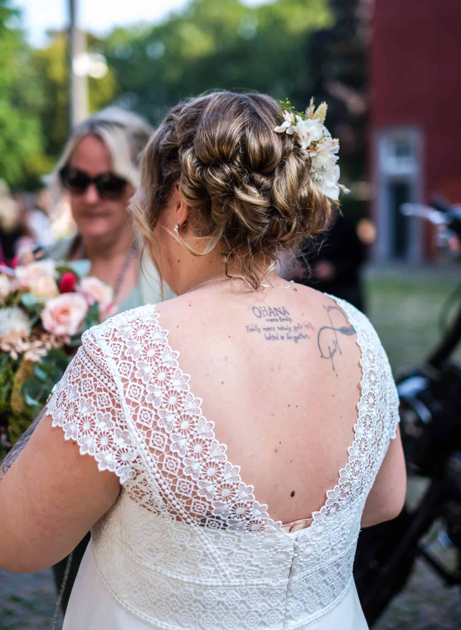 Hochzeitsbraut mit verspielt geschlungener Hochsteckfrisur und Blumenaccessoire, Blick auf Tattoo am Rücken.