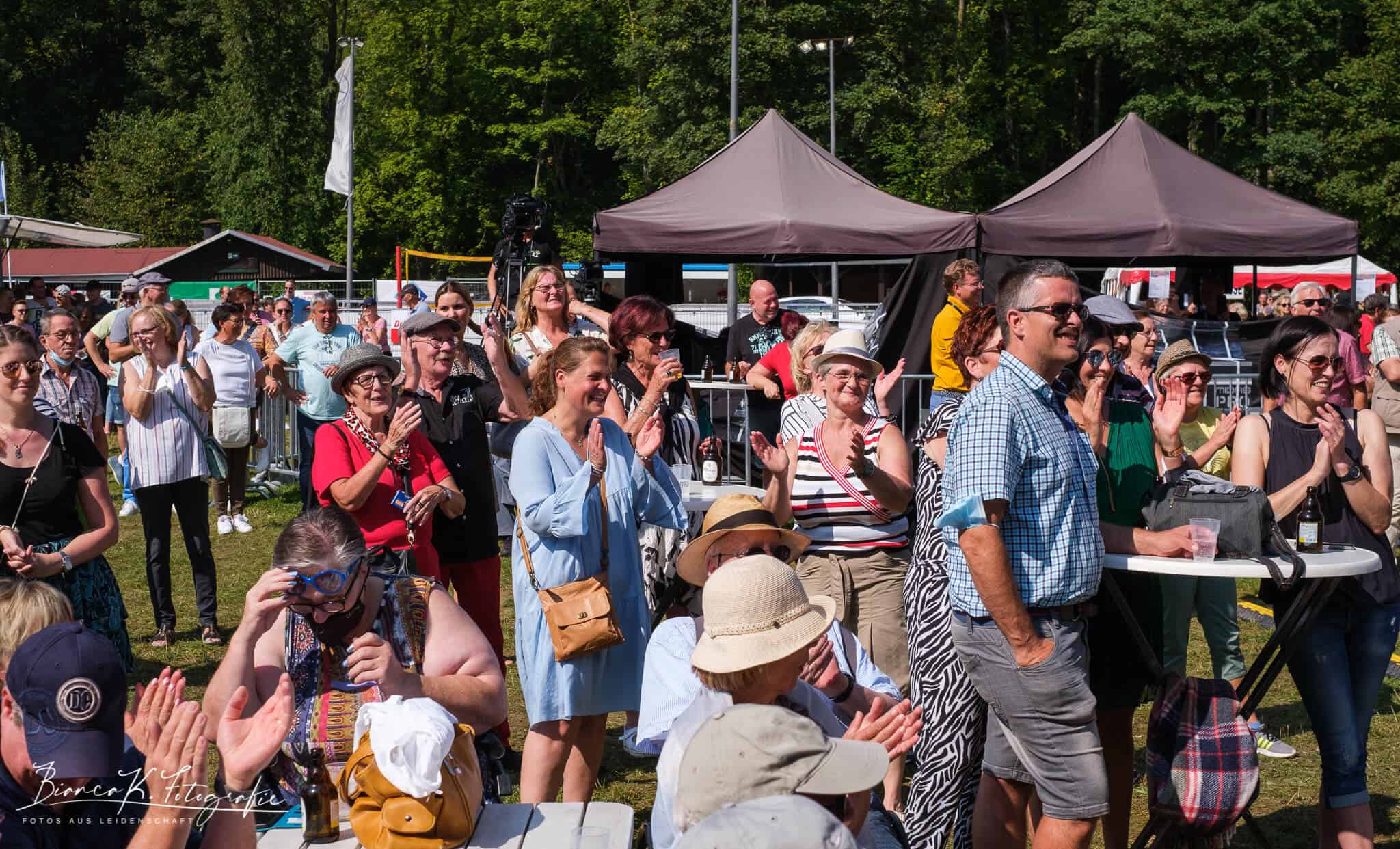 Lebensfrohe Menschen beim Open-Air-Festival im Grünen, lächelnd, klatschend und fröhlich feiern bei Sonnenschein.