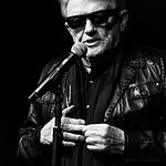 Stimmungsvolles Schwarz-Weiß-Foto eines älteren Mannes mit Sonnenbrille, Mikrofon und Lederjacke bei einer Rede.