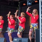 Fröhliche Frauen singen und tanzen bei einem Konzert auf der Bühne, tragen rote Hemden und bunte Shorts.