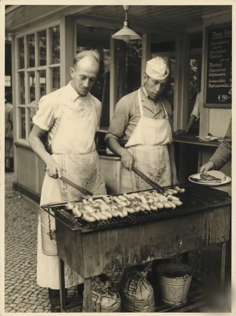 Zwei Männer in Rüter-Schürzen grillen Würstchen über einem langen Holzkohlerost vor dem Verkaufsstand