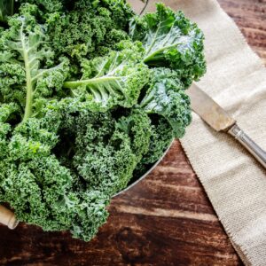 Frischer regionaler Grünkohl aus Niedersachsen mit saftig-grünen Blättern, ansprechend drapiert in einem Metallsieb mit Holzgriffen auf einem rustikalen Holzuntergrund – pure Vorfreude auf die traditionelle norddeutsche Winterküche.