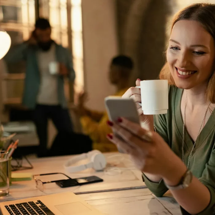 Junge Frau blickt lächelnd auf ihr Smartphone, in der anderen Hand eine Kaffeetasse – an einem Arbeitsplatz mit Laptop in warmem Licht