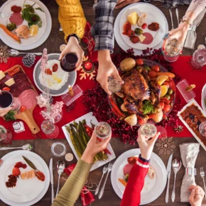 Weihnachtlich gedeckter Tisch von oben mit gebratenem Geflügel in der Mitte, umgeben von Tellern mit Aufschnitt, Lachs, Spargel und Salat; mehrere Personen stoßen mit Wein- und Sektgläsern über der Festtafel an