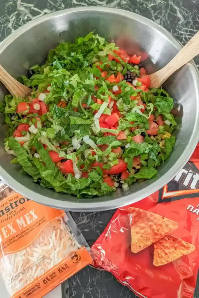 adding the lettuce to the top of the salad in large salad bowl. Packages of shredded cheese and nacho Doritos sit on either side.