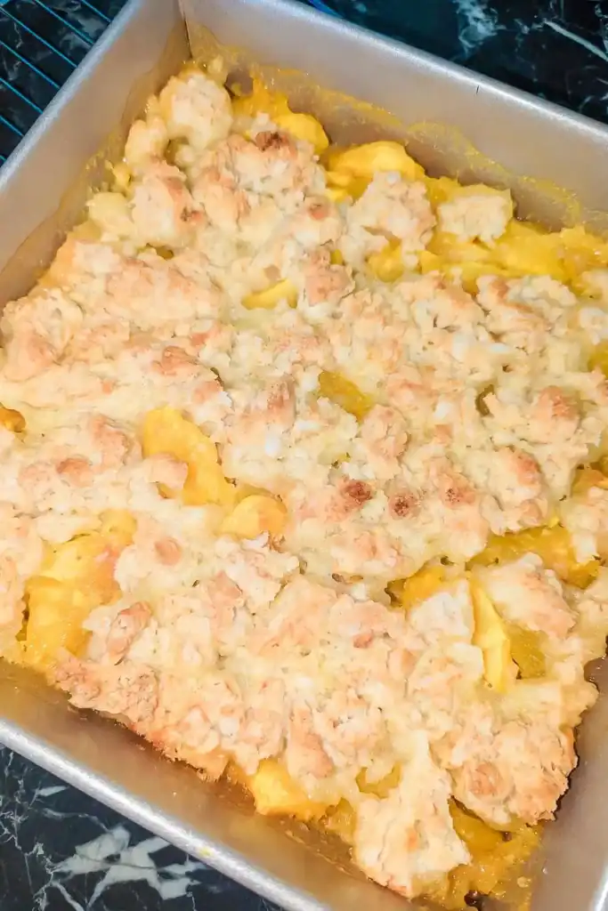 the baked peach cobbler in a light grey non-stick baking pan on wire rack on black marble table