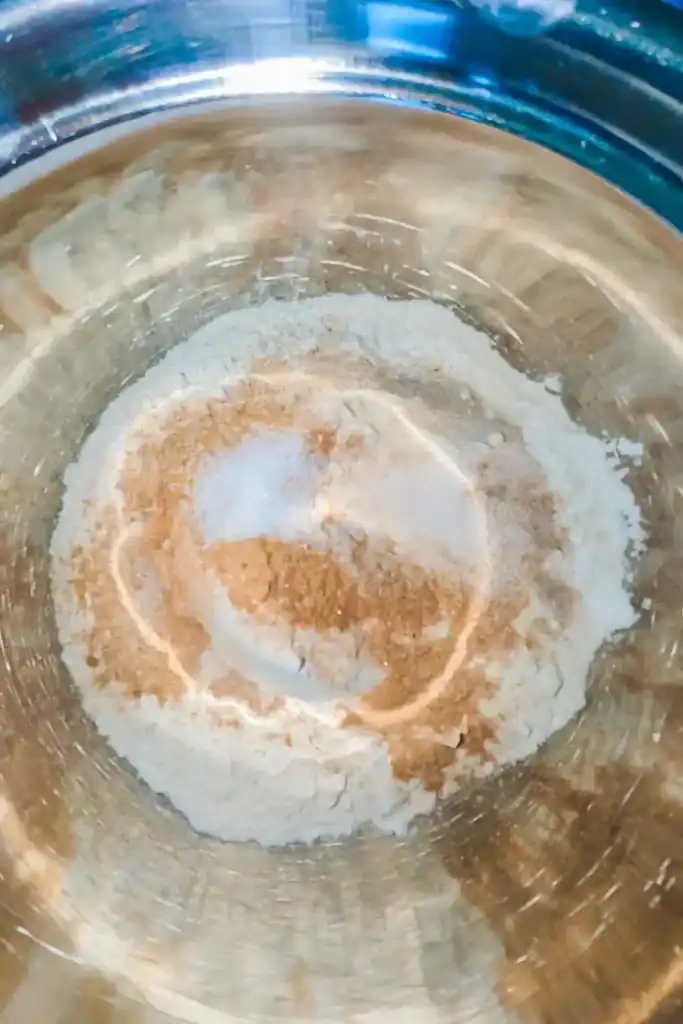 adding cinnamon and sugar to dry ingredients in stainless steel mixing bowl